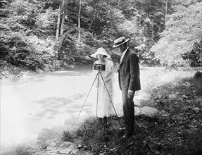Woman with camera and man in woodland setting, next to a creek  ca.  between 1910 and 1920