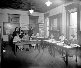 Women typists in an office, "Everybody Hustle" sign in the background ca.  between 1910 and 1935