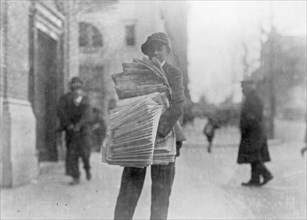 Newsboy on a city street holding a stack of newspapers ca. between 1909 and 1920