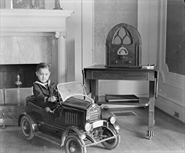 Child seated in toy automobile, radio on a table ca. between 1909 and 1923