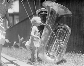 A toddler blowing on a tuba or euphonium ca. between 1909 and 1923
