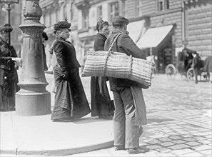 Man standing on a street corner in an Austrian city carrying baskets used to deliver meats and groceries ca. between 1909 and 1920