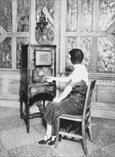 Woman, full-length, seated, facing left, turning dial of a console radio ca. 1909