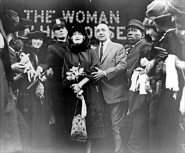 Mildred Harris Chaplin, full-length portrait, standing in crowd, facing front, guarded by a policeman (1920)