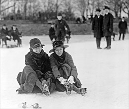 Two people seated on snow, and putting on ice skates, in the Washington, D.C., area ca. 1909