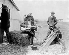 Two Afro-American men outside a stable, with Kazoos in their mouths, one of them seated on haybale and playing guitar, the other playing homemade string instrument ca. 1909
