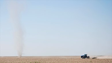 Farmer plowing a field on the Eastern Colorado plains, tractor facing left and driving toward a natural phenomena called a dust devil