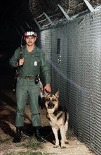 A US Army military policeman patrols a fence line with a patrol dog.