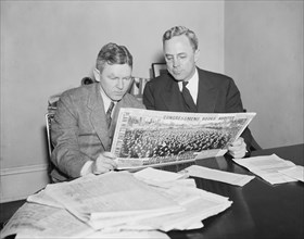 Two men sitting at a desk reading a newspaper