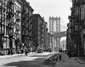 Looking down Pike Street toward the Manhattan Bridge, street half in shadow, rubble in gutters, some traffic. Pike and Henry Streets, Manhattan. ca. 1936