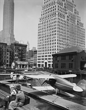 Two shoe-shine boys sit on edge of pier overlooking skyport, where seaplanes are moored; Manhattan office buildings beyond ca. 1936