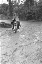 Dutch soldier on a motorcycle, dressed in a rain cape, drives on a muddy road. Barbed wire rolls lie in the bend; Date January 1947; Location Indonesia, Dutch East Indies, Palembang, Sumatra