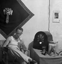 Man in a room smokes a pipe while reading. On the right a table with radio and books. Behind him a tapestry depicting a barong - Indonesia, Dutch East Indies ca. 1947