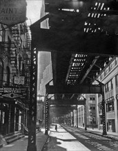 1930s New York City - Woman pauses below elevated train tracks, office buildings visible at left of tracks, painters shop in older building at left ca. 1936