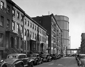 New York City Street lined with cars, houses with decorative ironwork in foreground, apartments, large gas tanks beyond ca. 1938