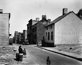 African American woman sits at street edge with two children, empty lots on either side of street, old 2 and 3 story clapboard houses further up. Talman Street, between Jay and Bridge street, Brooklyn...