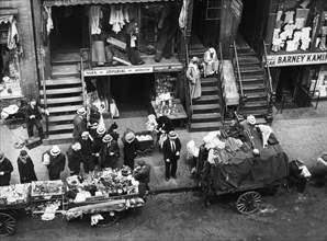 Looking down from window on New York City street scene including peddlers, women sitting on stoop, baby carriages, fabric and clothing stores and pedestrians. ca. 1938