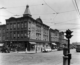 Clapboard buildings, cobblestone streets with trolley tracks, cars, including a taxi and an entrance to the subway on the corner. Graham and Metropolitan Avenues, Brooklyn. ca. 1937