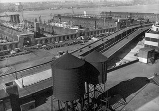 Eight cars visible on Westside highway, beyond water towers in foreground, piers have entrances on street, ships visible at dock ca. 1937