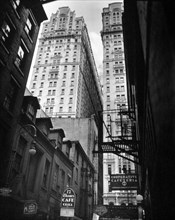 View of Thames Street, Manhattan New York City street scene. Low, older buildings, signs for bars, cooperative cafeteria, etc., street lamps and firsecapes in foreground, tall buildings in the light b...