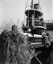 Tugboat Watuppa moored at Pier 5, bow of boat with rope bumpers in foreground, crew posing on board, Manhattan in the distance ca. 1936