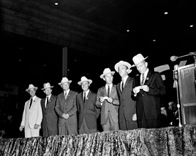 The original seven Mercury astronauts, each wearing new cowboy hats and a badge in the shape of a star, are pictured on stage at the Sam Houston Coliseum in Houston, Texas.