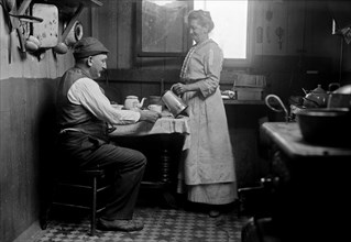 Woman pouring a drink for a man seated at a table in a house in Broad Channel, Queens ca. 1910-1915
