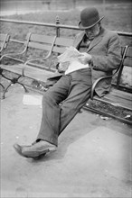 Man sitting on a park bench in Union Square, reading a newspaper ca. 1910-1915