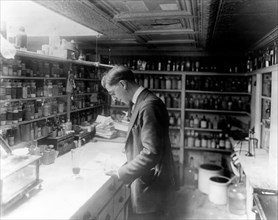 Pharmacist at People's Drug Store, No. 5, 8th and H Streets, N.E., Washington, D.C., looking at prescriptions(?) on the counter in room lined with shelves of pharmacy bottles ca. 1909-1932