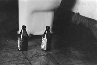 Two bottles of beer on a tile floor near a toilet bowl - Brand: E&B Light Lager Beer; Date 1947; Location Indonesia, Dutch East Indies