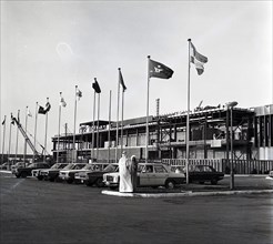 A photograph of the exterior of Bahrain International Airport in December 1975. Parked taxis can be seen in the foreground amongst flag poles