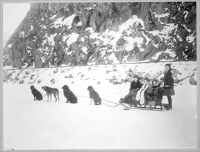 Dog team harnessed to a sled holding four girls, in front of a snow-covered cliff. 1900-1917 Canada