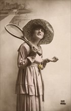 Woman in dress and hat with tennis racquet and ball.