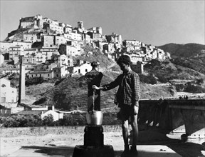 europa, italie, calabre, corigliano calabro, garçon à la fontaine avec le village en arrière-plan, 1962