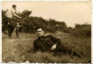 Soviet Army captain lying in the grass on the banks of the Elbe River in the background a man riding a bicycle on a footpath.