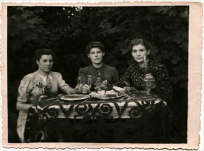 Three young women holding glasses of alcohol, sitting at a table covered with in garden.
