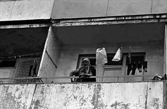 An old woman standing on the balcony of a multi-storey panel house.