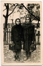 Two school girls with briefcases in their hands are against the fence.
