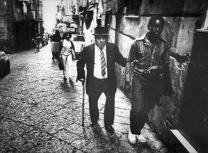 Elderly man and black man walking in a street of naples, 80's