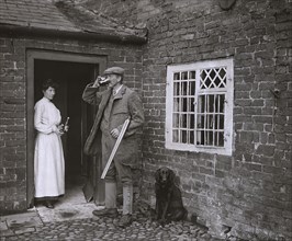 A man with a gun drinking beer and a lady in the doorway of an old cottage holding bottle and waiting for glass.