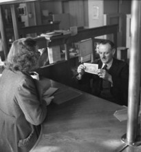 Client at post office counter; post office official checking banknote for its authenticity;  1943.