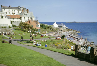 People sittng on grass by the coast enjoying sunshine at the seaside resort of Portrush, County