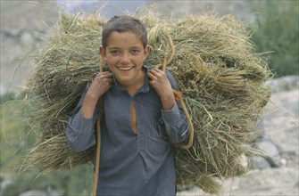 PAKISTAN, Hunza, Altit, Smiling boy carrying load of hay on his shoulders