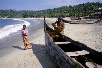 INDIA, Kerala, Kovalam, Fisherman tending to his boat moored on the sandy beach with passing man in