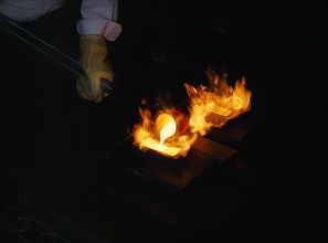 INDUSTRY, Factory, Gold Manufacturing, Gloved hand holding pole pouring molten gold into flaming