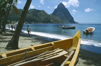 WEST INDIES, St Lucia, Soufriere, People walking past fishing boats on beach with the Pitons in the