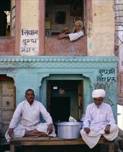 INDIA, Rajasthan, Bikaner, Two men sitting on table outside shop with man framed  in window above.