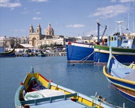 MALTA, Marsaxlokk, The Harbour with the prows of colourful moored fishing boats with the cathedral
