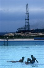 CUBA, Tibacoa Resort , Two men sitting in a shallow swimming pool with Oil derrick in the