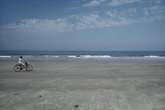 INDIA, Goa, Colva Beach , Cyclist riding along sand of the empty beach with waves rolling on to the